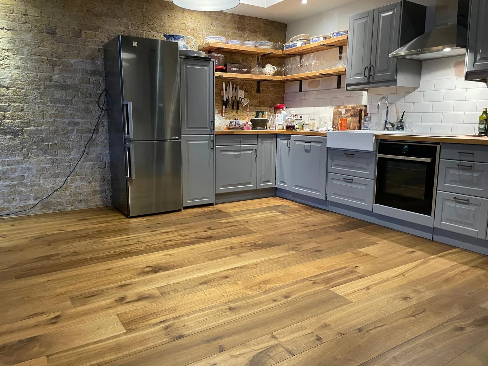 Grey shaker kitchen with oak shelves and a rustic oak floor against exposed brick